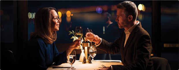 A couple toasting with wine glasses at a romantic dinner in The Point Revolving Restaurant, with dim lighting and city lights in the background.