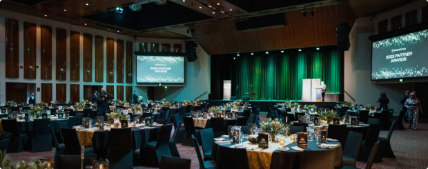 Spacious event hall set up for a gala with round tables dressed in linens, a large screen displaying event details and a stage.