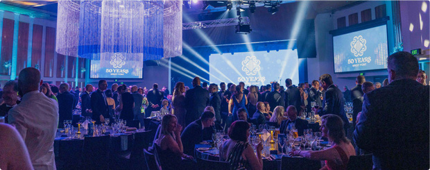 Elegant gala event scene with guests in formal attire, dining under blue lights and large chandeliers.
