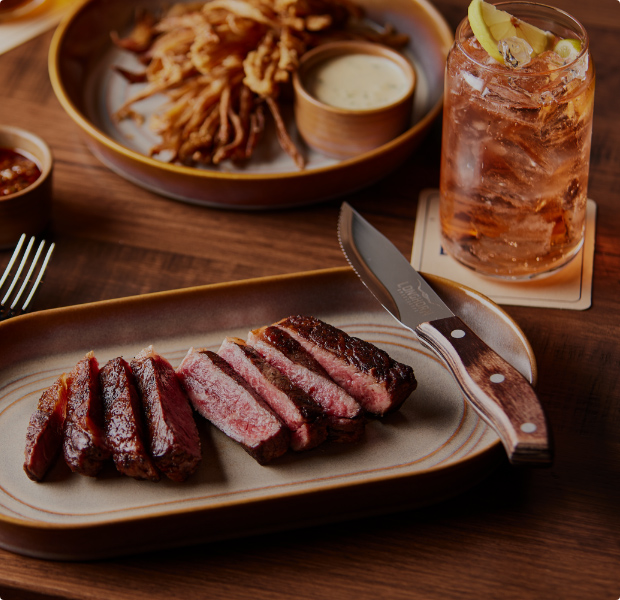 Savor the rich flavors of a perfectly cooked steak served with golden onion rings and a refreshing drink at Longhorn Smokehouse.