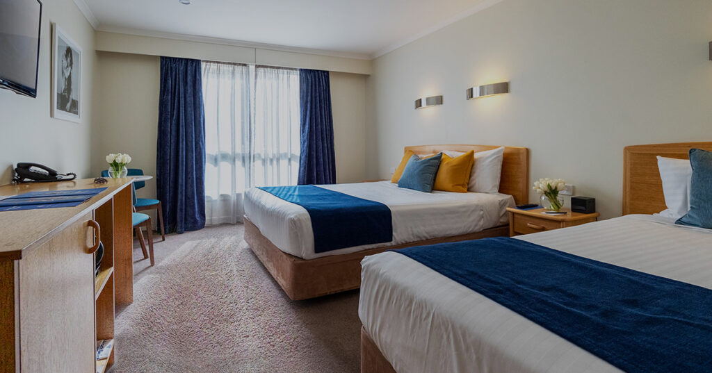 Comfortable twin room at Wrest Point's Motor Inn featuring two plush beds with blue runners, wooden furniture and ample natural light from large windows.